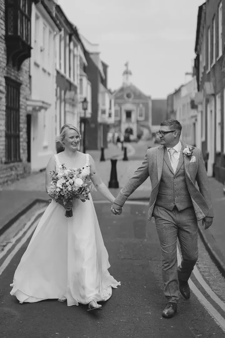Black and white portrait of bride and groom walking hand in hand through Poole Old Town with the Guildhall at the end of the street