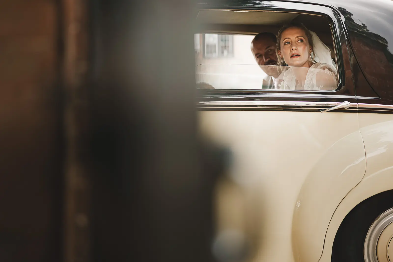 Candid documentary portrait of a veiled bride looking out the window of a cream vintage wedding car with quiet anticipation, framed through a foreground architectural element