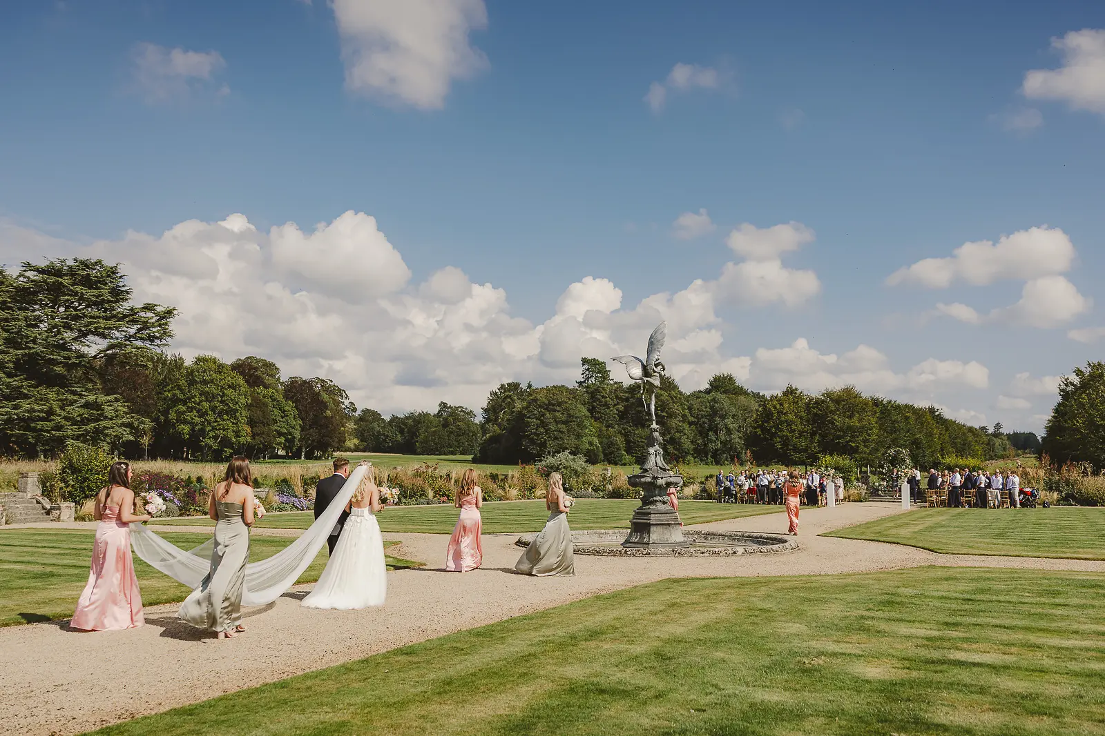Bride and bridesmaids processing through the formal gardens of St Giles House Dorset on a sunny summer day, bridesmaids carrying the bride's long train past an ornate fountain