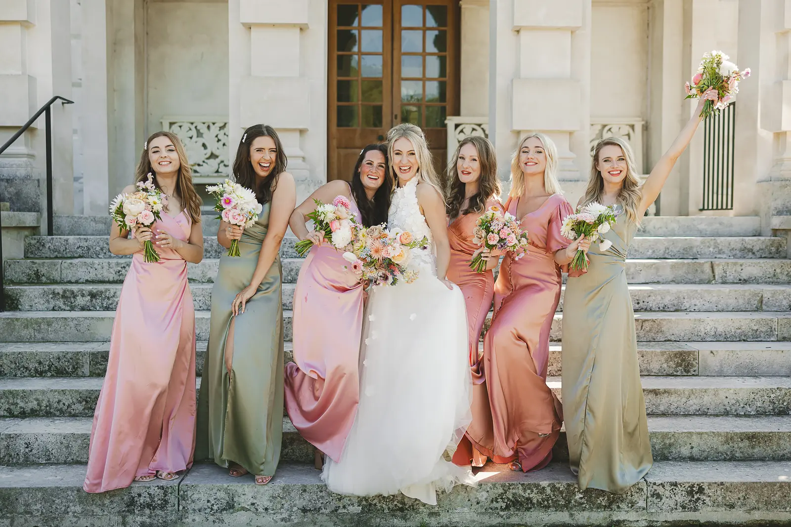 Bride and five bridesmaids laughing and celebrating on the grand stone steps of St Giles House Dorset, bridesmaids in mismatched satin gowns in dusky pink, sage and terracotta