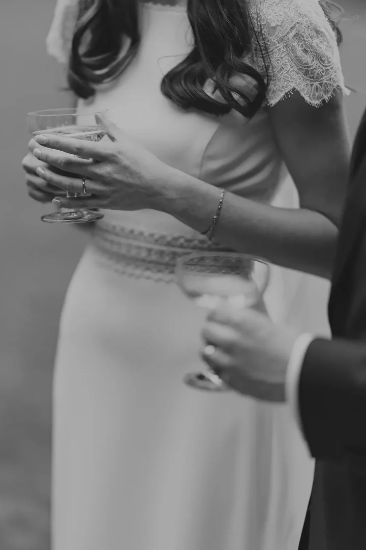 Black and white close-up of bride holding a champagne coupe glass with groom's arm around her, showing lace cap sleeve dress and delicate diamond bracelet