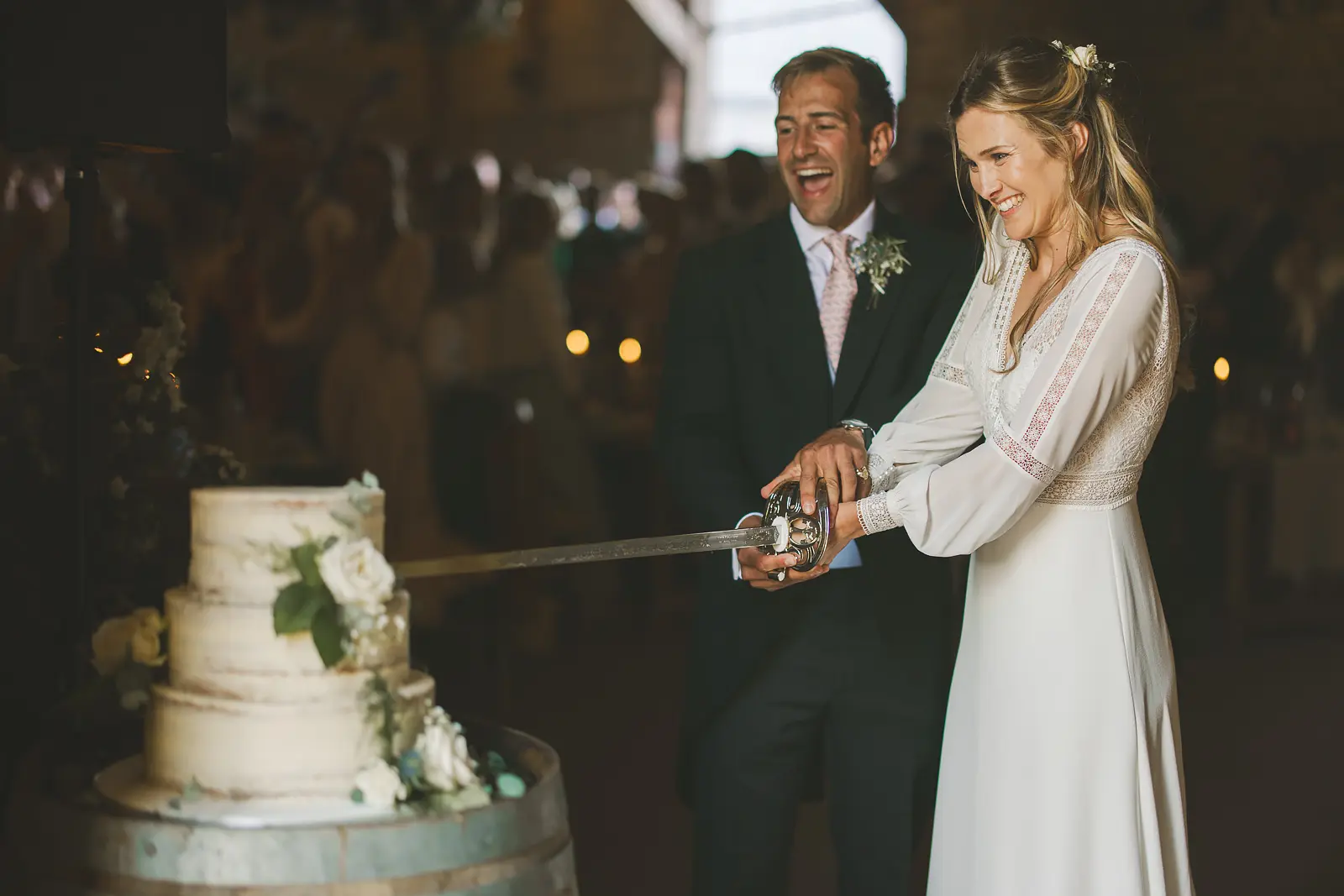 Bride in a boho lace gown wielding a ceremonial sword to cut a three-tier naked wedding cake on a wine barrel at a candlelit barn reception while her groom laughs behind her