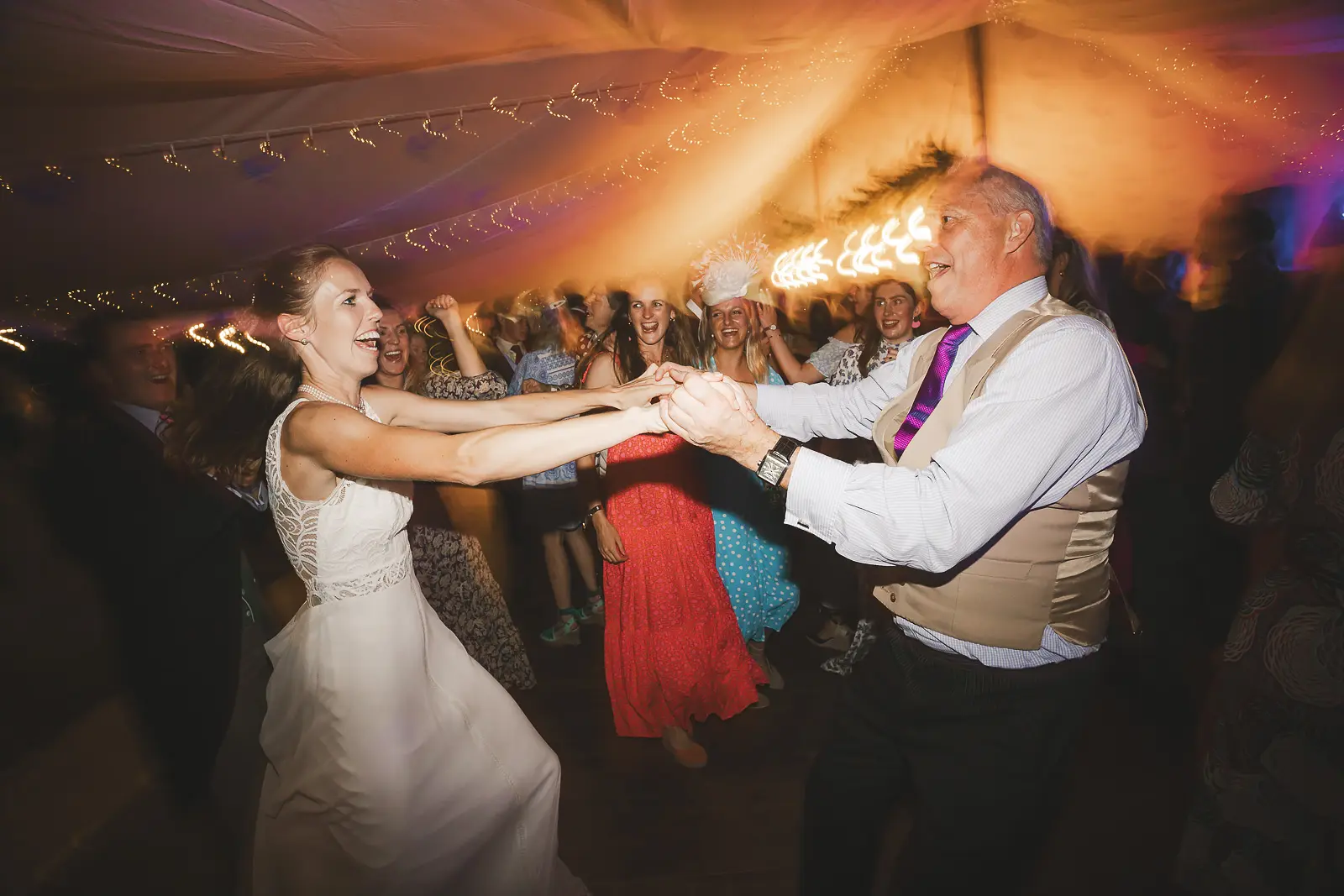 Bride in a lace gown dancing energetically with her father on the marquee dance floor as cheering guests crowd around them under colourful evening reception lights