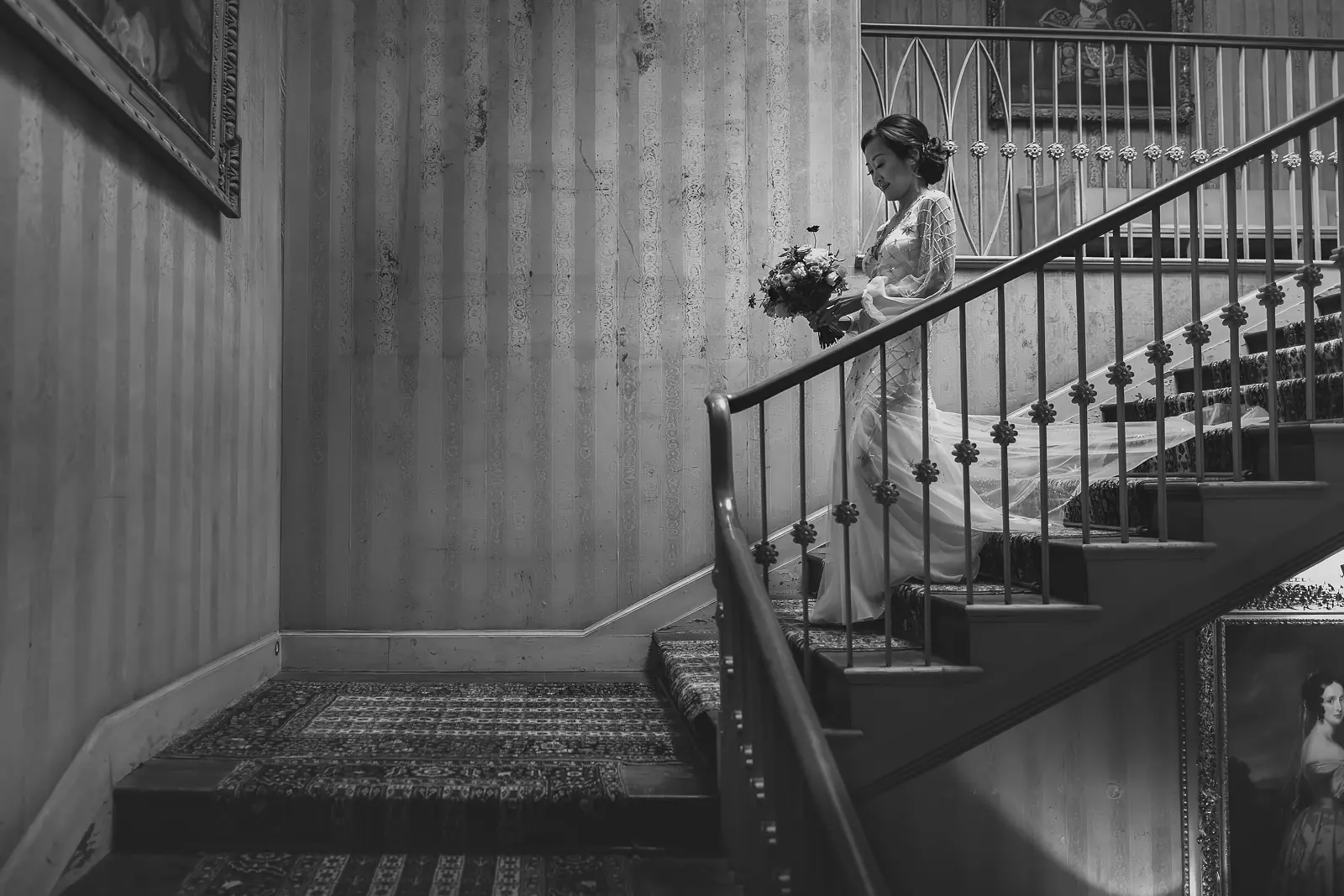 Black and white portrait of a bride descending the curved staircase at St Giles House, Dorset, holding a wildflower bouquet with antique portraits lining the striped walls