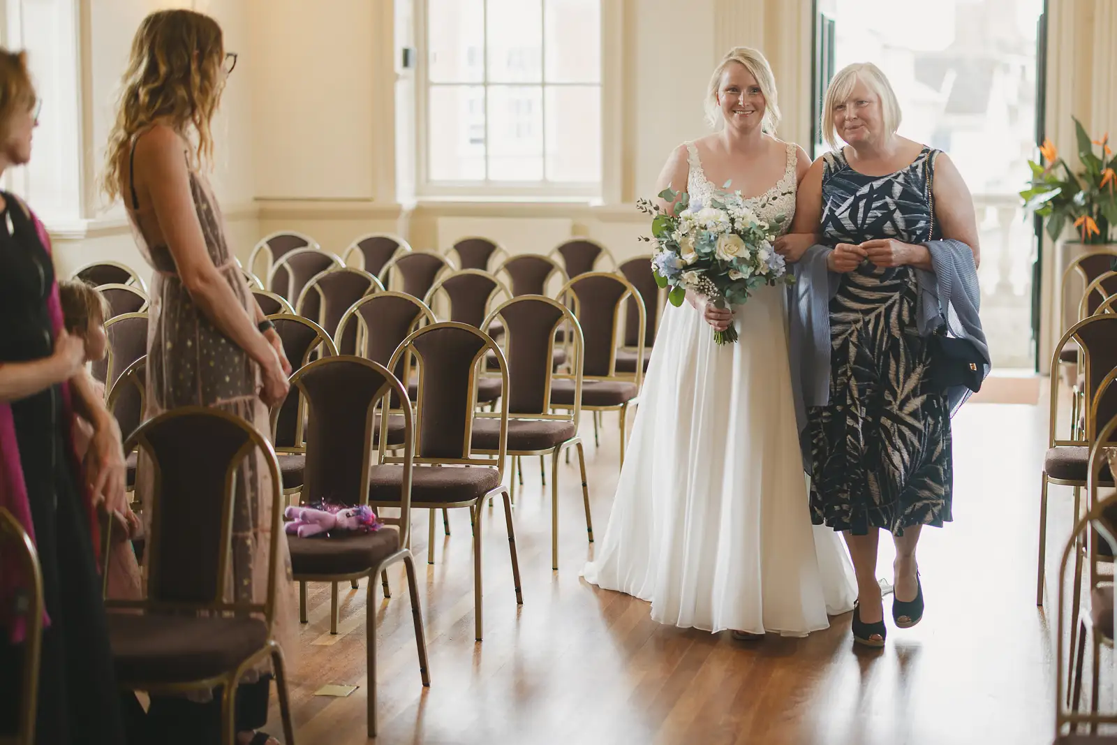 Bride smiling as she walks down the aisle with her mother inside Poole Guildhall ceremony room, blue and white bouquet in hand