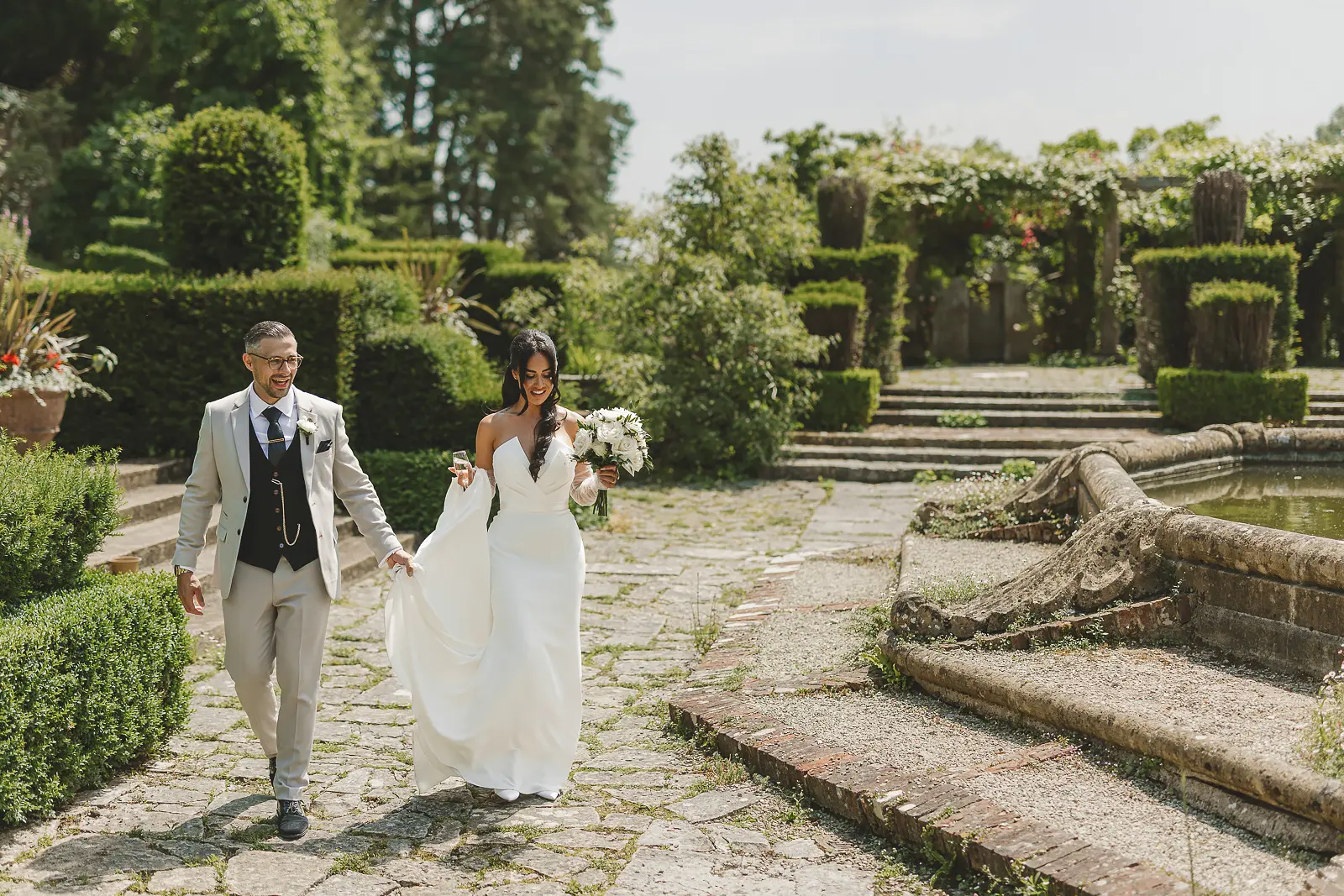 Bride laughing with her father as they walk through a formal garden with clipped box hedges and a stone fountain, bride in a strapless fitted gown with white bouquet