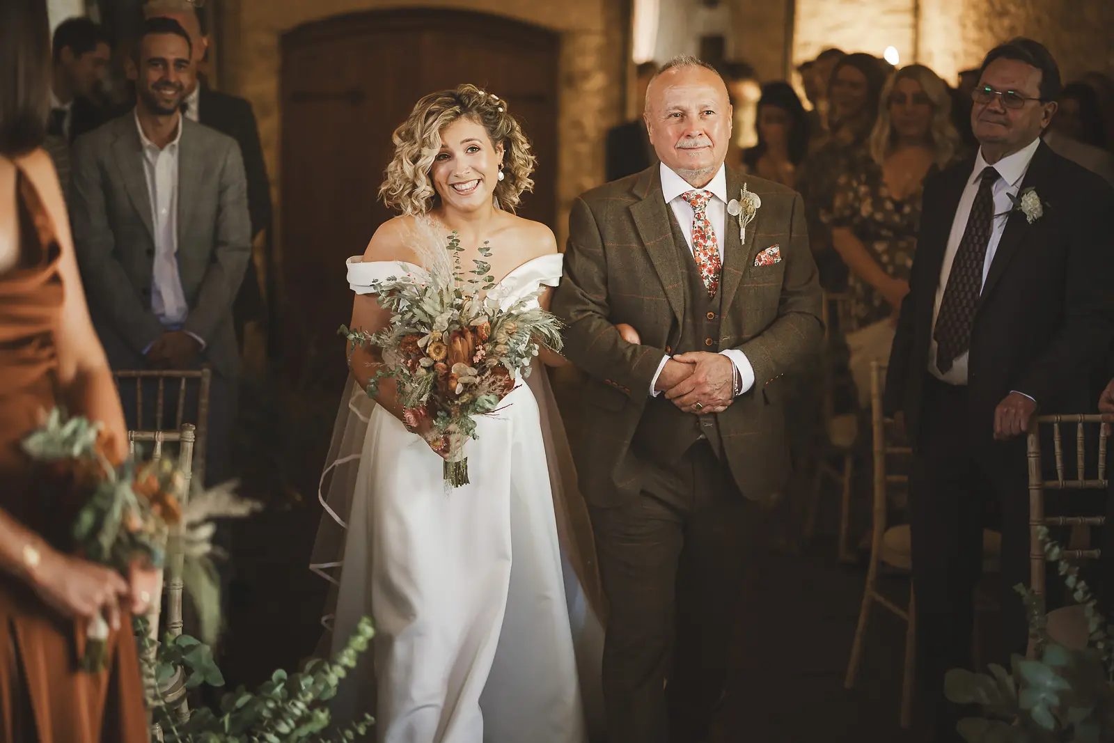 Radiant bride with curly hair walking down the aisle arm in arm with her father, carrying an autumn dried flower and pampas grass bouquet in a warmly lit stone barn ceremony venue