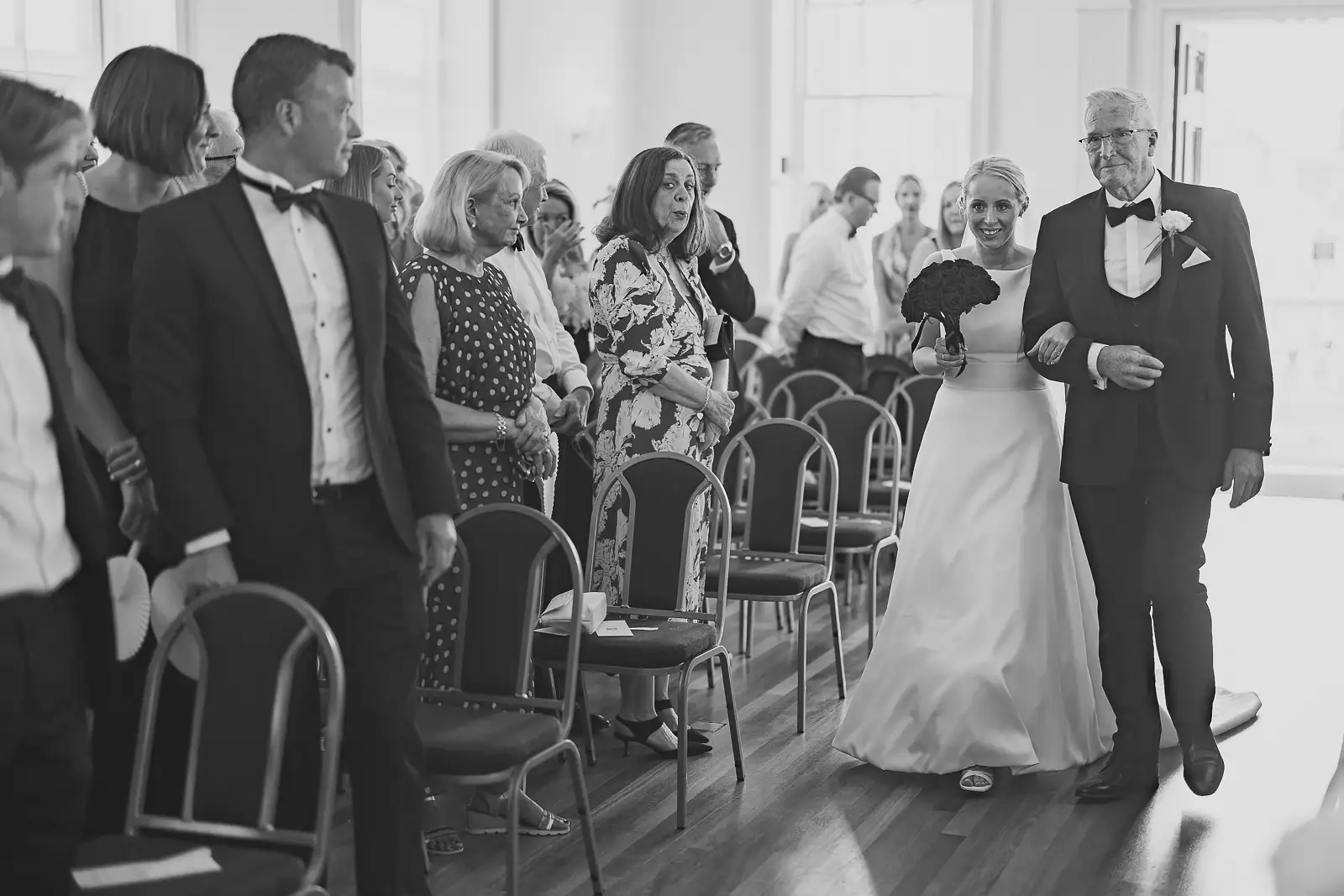 Black and white photograph of bride with dark bouquet walking down the aisle with her father as guests stand at Poole registry office wedding