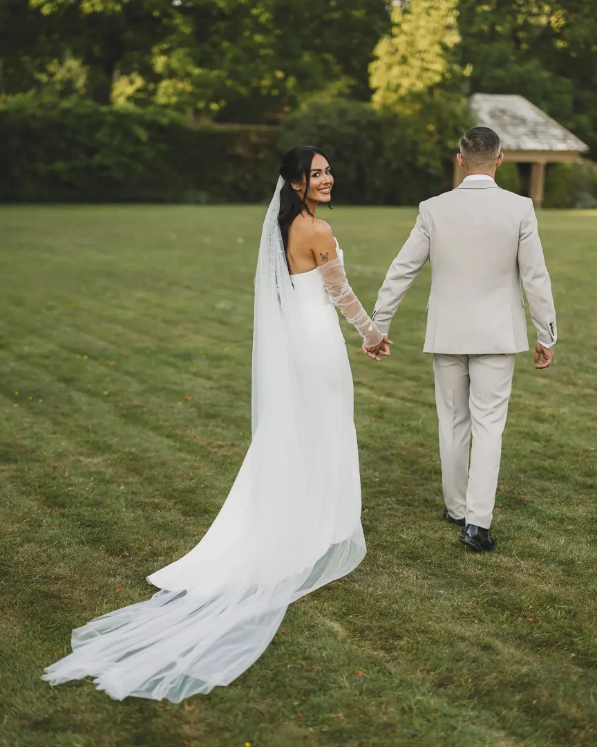 Bride glancing back over her shoulder smiling while walking hand-in-hand with groom across a manicured lawn, in a backless gown with cathedral train and one lace sleeve