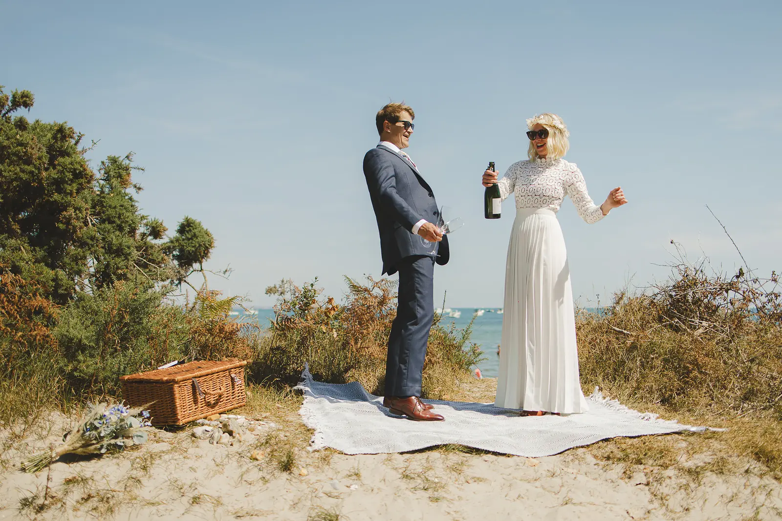 Bride and groom in sunglasses enjoying a champagne picnic on the sand dunes of the Dorset coast with a wicker hamper, blue sea visible behind them
