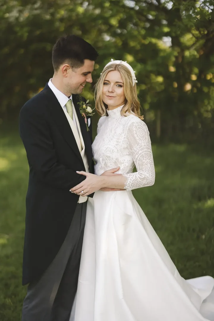 Elegant wedding portrait of bride in a high-neck lace bodice gown and pearl headband with groom in morning suit, bathed in warm golden evening light against a lush garden
