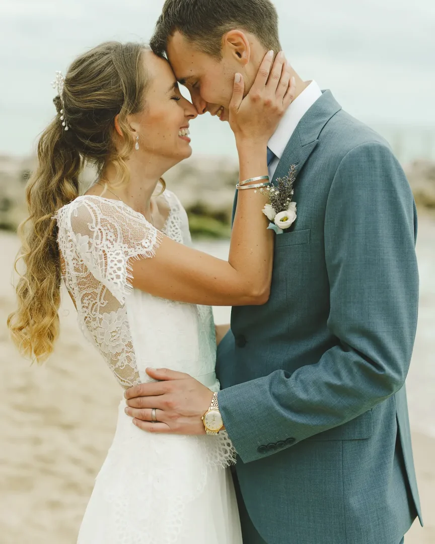 Intimate wedding portrait of bride in lace dress and groom in blue-grey suit touching foreheads and smiling on a sandy beach with rocky coastline behind them
