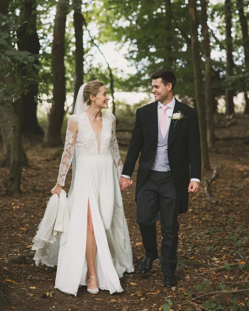 Bride and groom walking hand in hand along a dappled woodland path, bride in a long lace deep V-neck gown and groom in morning suit with pink tie, smiling at each other