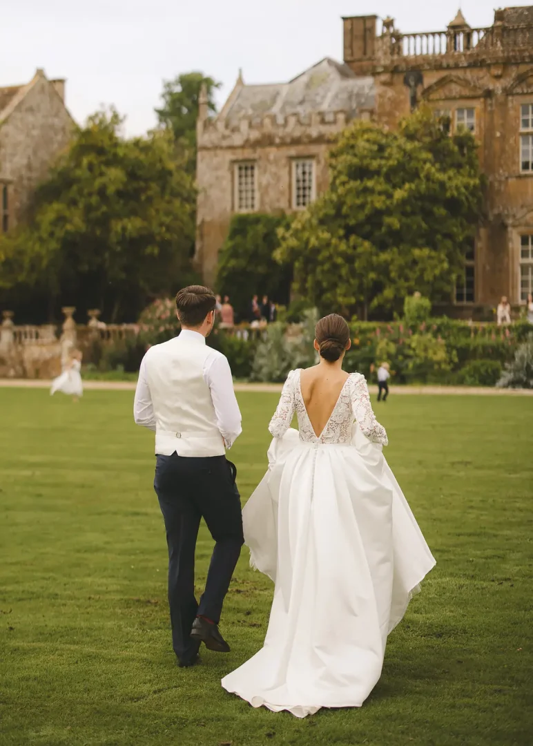 Bride in a deep V-back lace gown and groom in morning suit walking across the lawn toward Mapperton House, the Jacobean manor surrounded by formal gardens in Dorset