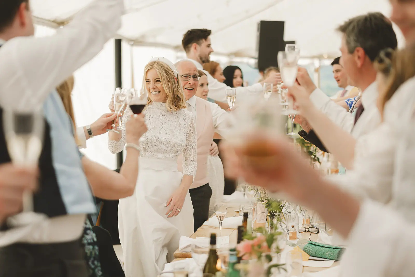 Beaming bride in a high-neck lace dress holding a glass of red wine as wedding guests raise their glasses in a joyful toast at a bright marquee reception