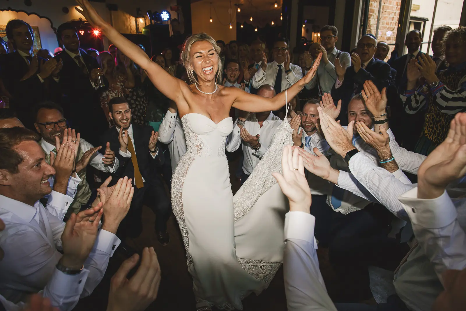 Bride lifted on a chair with arms raised wide in pure joy during the hora dance at a Jewish wedding at Larmer Tree Gardens Dorset, surrounded by ecstatic dancing guests
