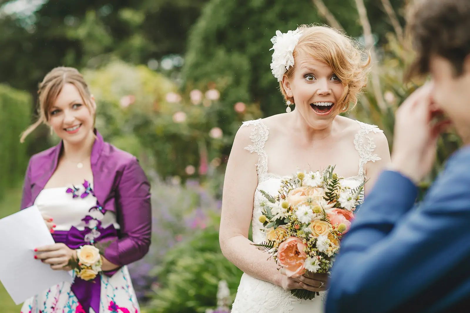 Bride with a wide joyful expression holding a peach and white bouquet during an outdoor garden ceremony, bridesmaid in purple smiling behind her