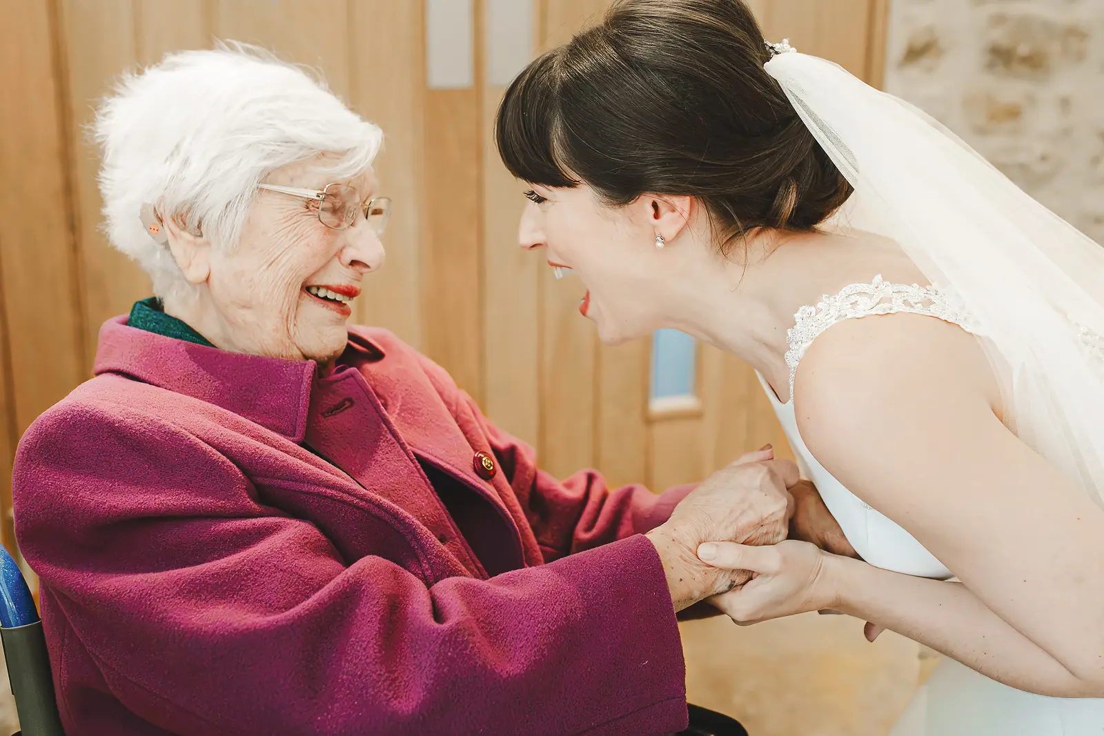 Bride bending down laughing and holding hands with her elderly grandmother seated in a chair at a Dorset wedding, a joyful and heartwarming documentary moment