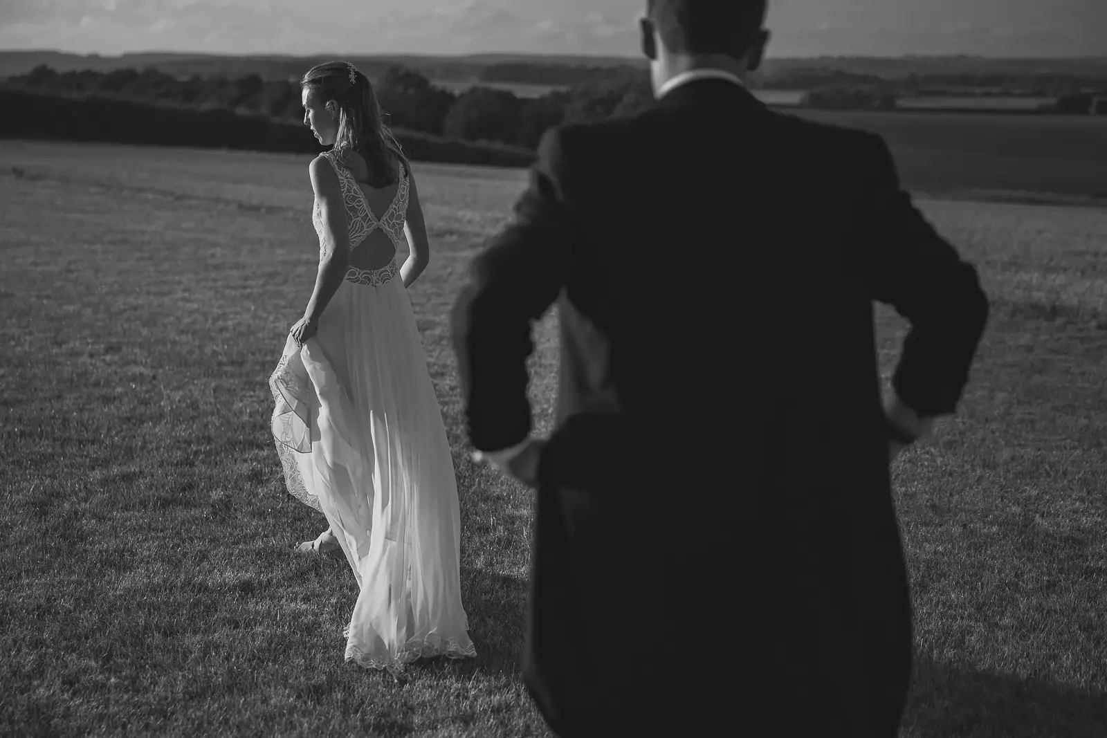 Cinematic black and white image of a bride in an open-back lace gown walking across a field as her groom watches, with the rolling Dorset countryside hills stretching behind