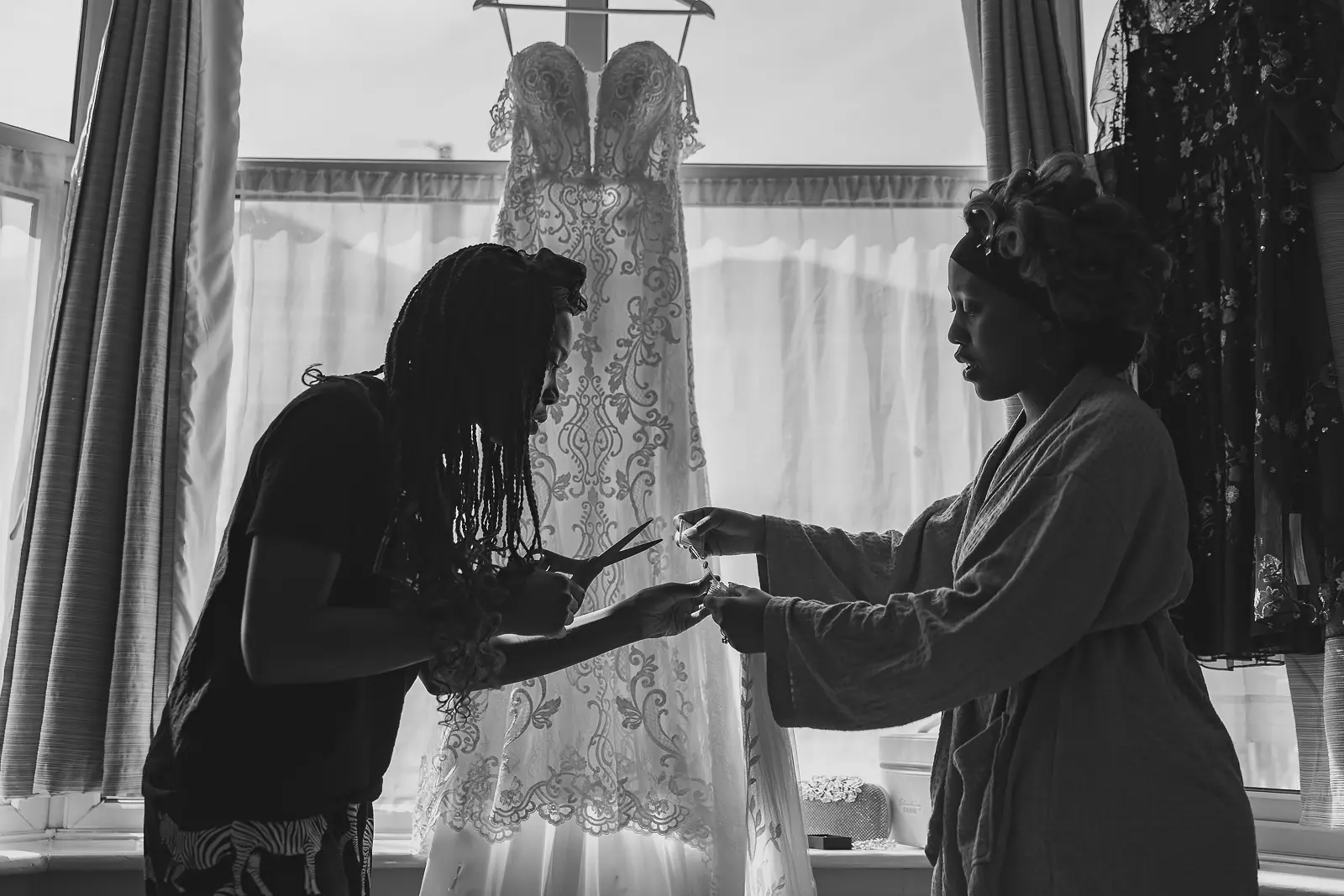Black and white silhouette of bride in robe and hair rollers with her stylist, an intricate lace wedding gown hanging backlit in the window behind them, Bournemouth