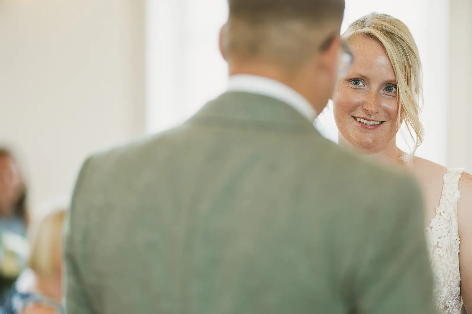 Bride smiling warmly at her groom over his shoulder during wedding ceremony at Poole Guildhall, lace dress in white light