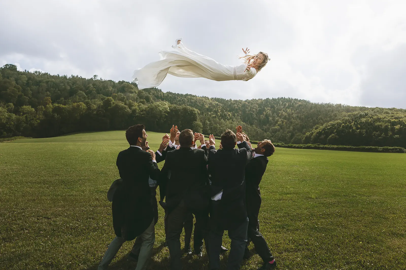 Bride being thrown high into the air by groomsmen in an open green field with wooded Dorset valley hills in the background, wedding dress billowing dramatically