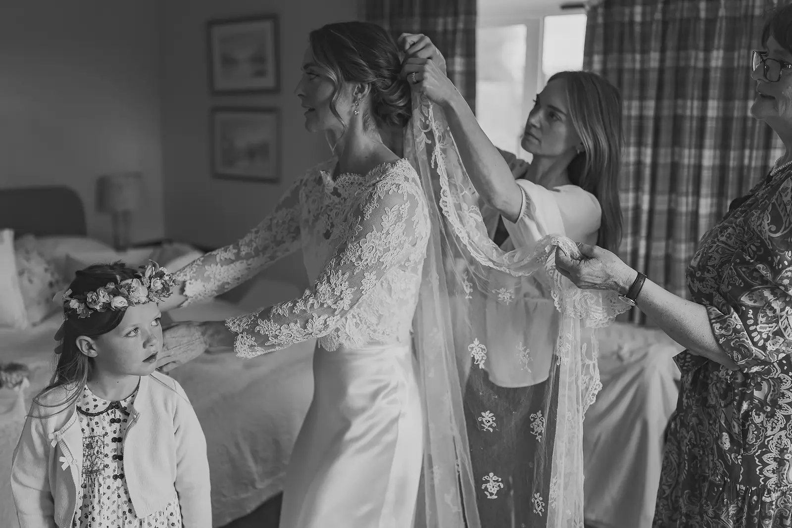 Black and white getting-ready scene of bride in a lace long-sleeve gown having her veil pinned while a little flower girl in a floral crown looks up watching adoringly