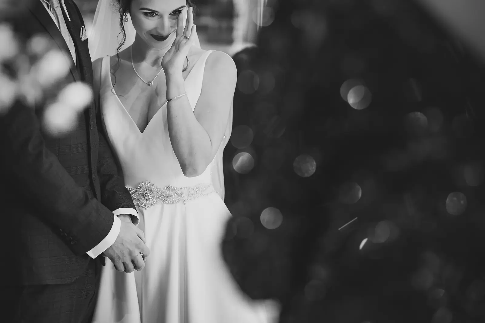 Black and white close-up of bride smiling through tears as she wipes her eye during the wedding ceremony, in a v-neck gown with a crystal beaded waistband, Dorset