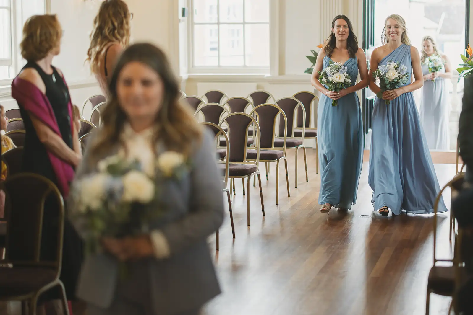 Bridesmaids in dusty blue dresses walking down the aisle carrying white bouquets inside Poole Guildhall ceremony room