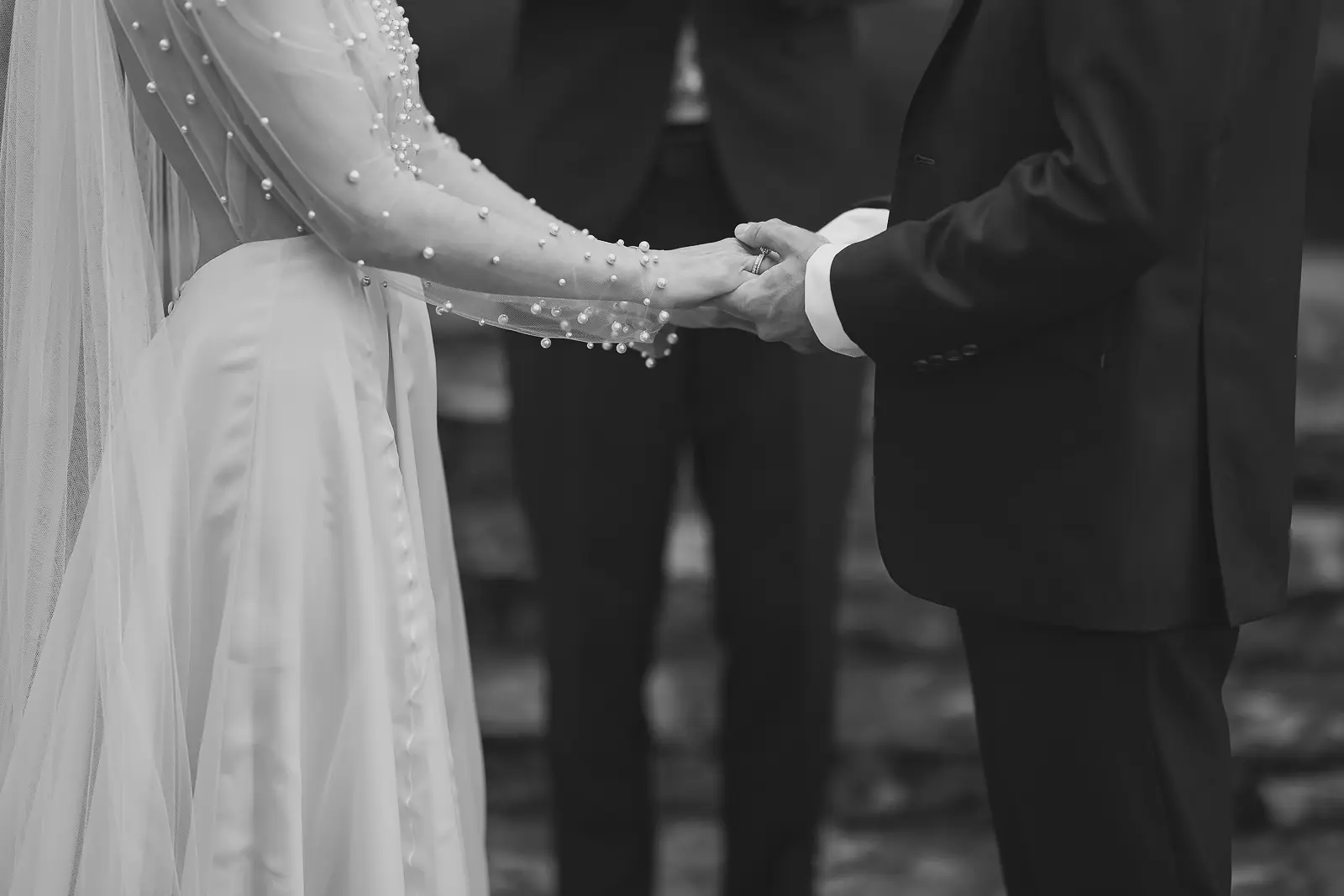 Black and white close-up of bride and groom holding hands during ceremony, bride in a pearl-embellished sheer sleeve gown, wedding ring visible