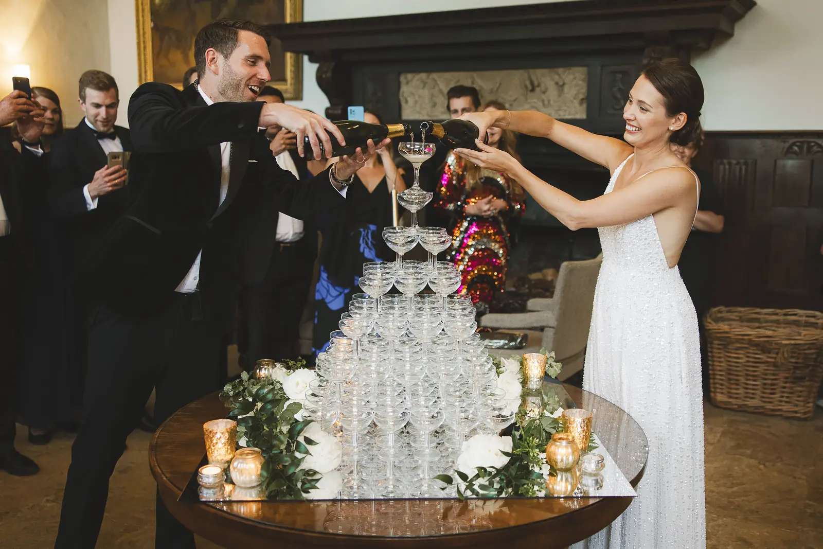 Bride and groom pouring champagne simultaneously onto a tall tower of coupe glasses at their Mapperton House wedding reception, guests watching in black tie around a stone fireplace