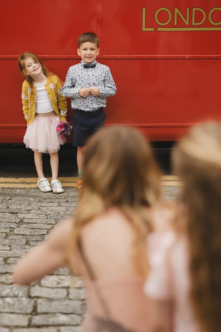 Young wedding guests in dressy outfits posing beside a red London bus on cobbled Poole street, girl in pink tutu