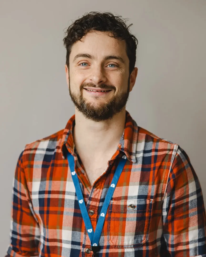 Natural commercial headshot of a smiling young professional in a plaid shirt against a clean grey background, Dorset