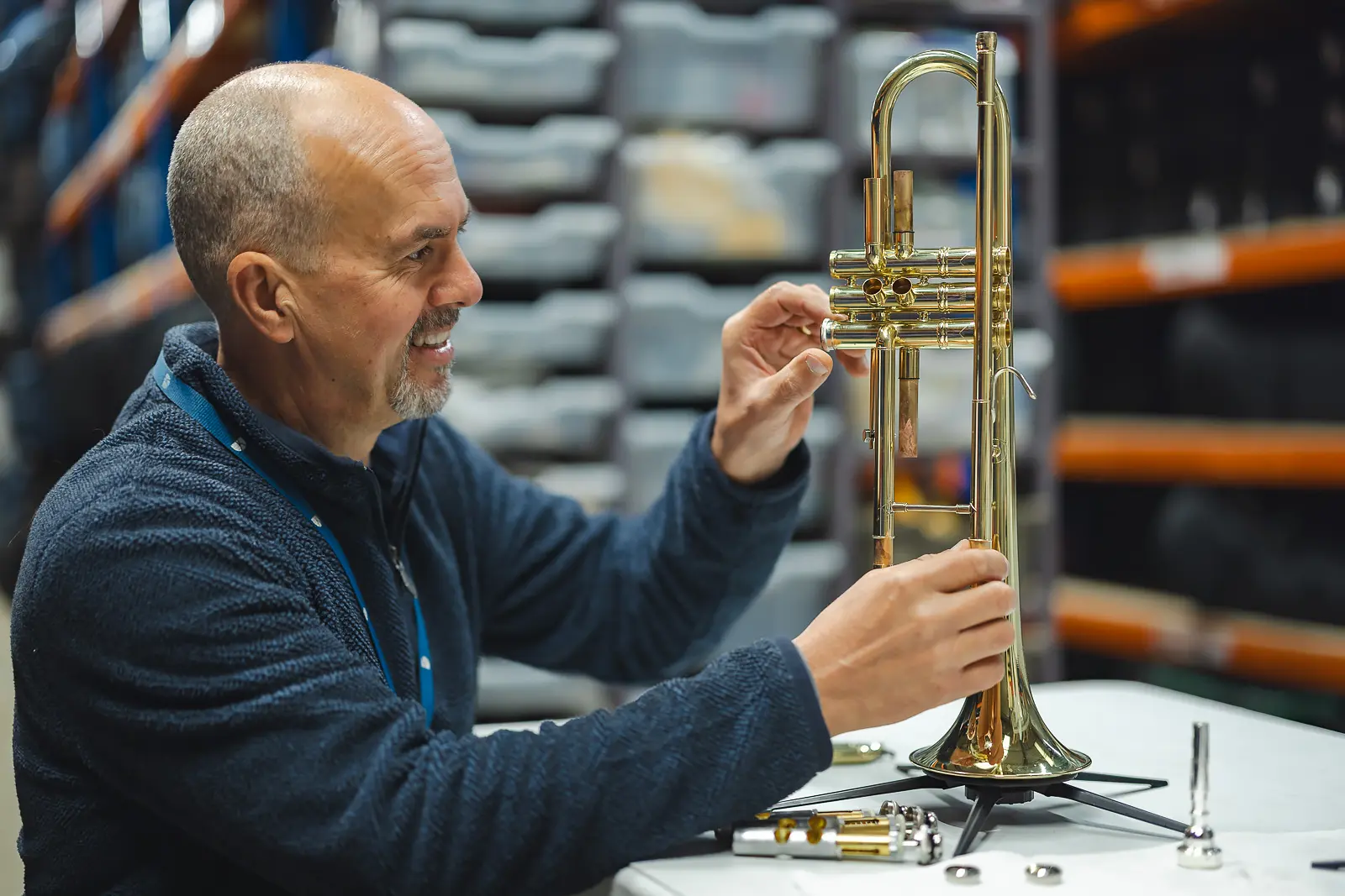 Instrument technician smiling while repairing a brass trumpet at his workbench in a music services warehouse in Blandford, Dorset