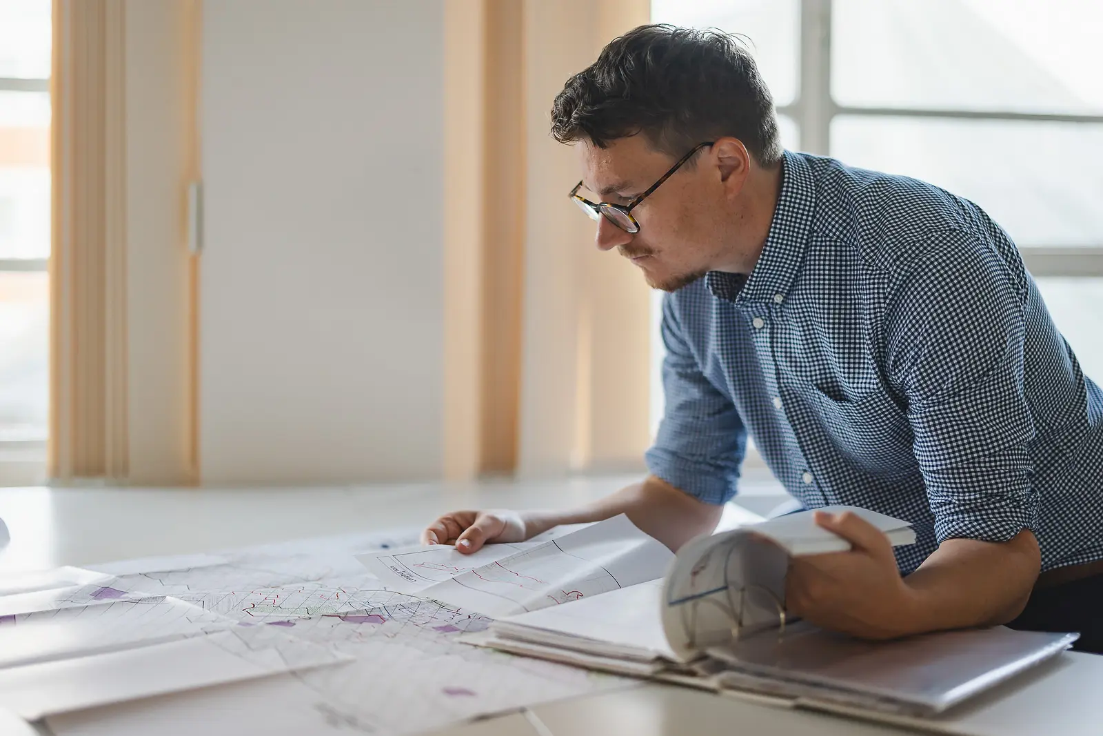 Young planning officer in glasses leaning intently over large planning maps spread across a desk in a bright Dorset office
