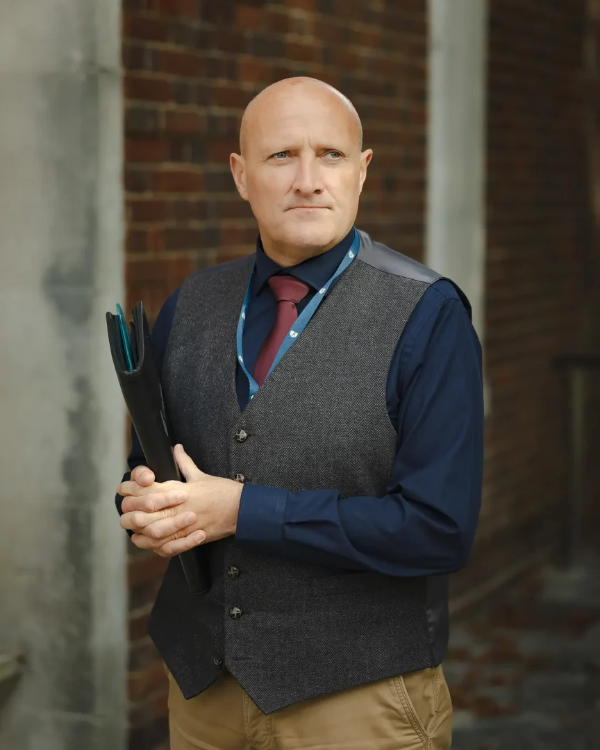 Senior professional in herringbone waistcoat and tie holding a folder, photographed against a brick wall in Dorchester