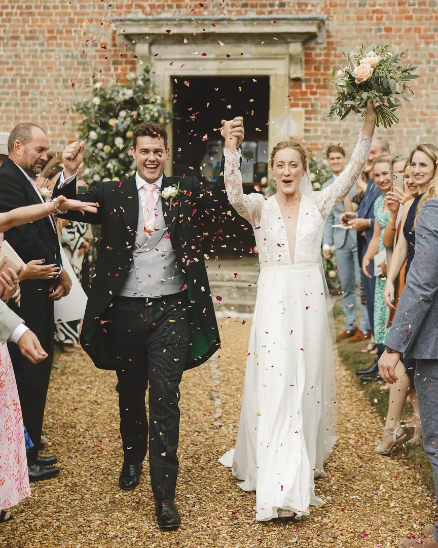 Bride raising her bouquet and groom lifting their arms with joy as guests shower them with colourful petal confetti outside the red brick doorway of a Georgian country house