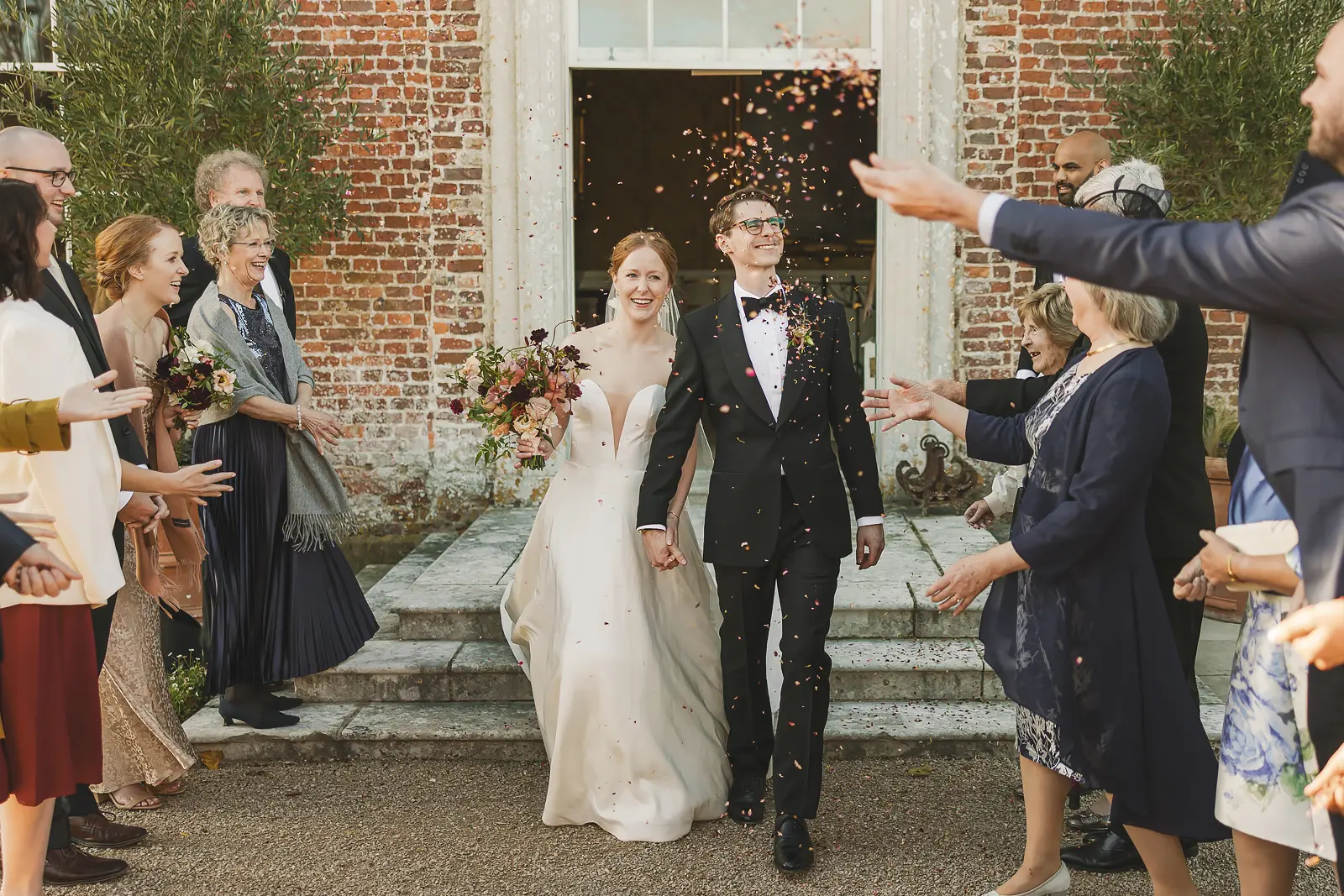 Bride and groom walking through a confetti shower on the steps of a Georgian red brick country manor, guests lining both sides throwing petals in autumn colours