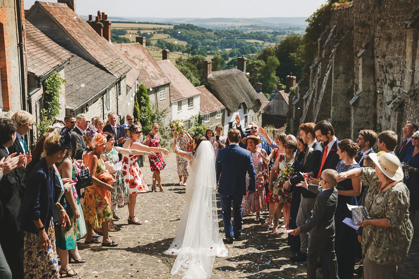 Bride and groom walking up the iconic cobbled Gold Hill in Shaftesbury as guests line both sides throwing confetti, rolling Dorset countryside stretching beyond