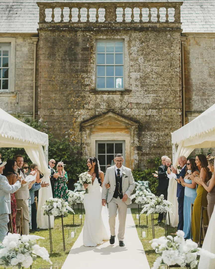 Bride and groom walking triumphantly down a white floral aisle at Mapperton House Dorset, the Jacobean stone manor providing a grand backdrop with guests applauding on either side