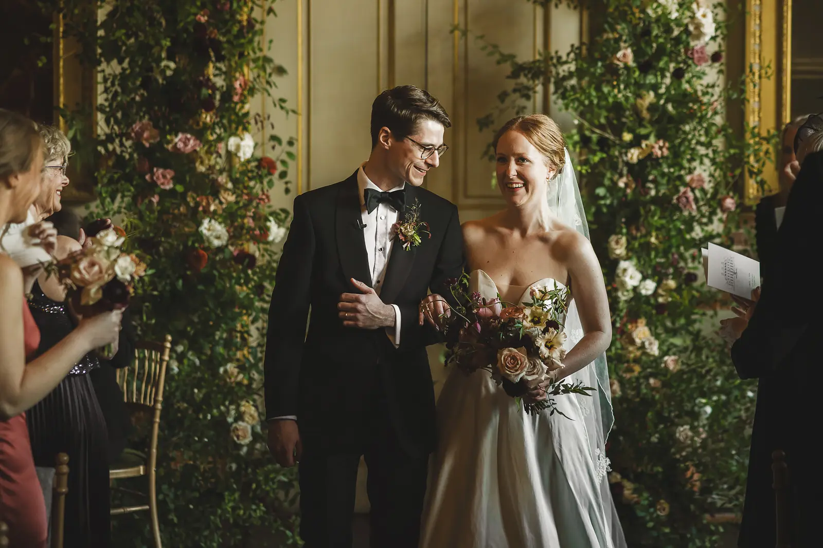 Bride and groom beaming as they walk out after their ceremony at St Giles House Dorset, flanked by spectacular floor-to-ceiling lush floral installations in the gilded state room