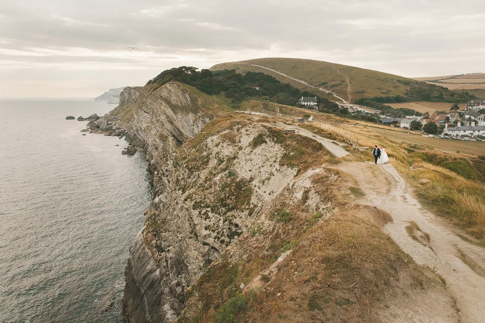 Bride and groom walking as tiny figures along a clifftop path above Lulworth Cove with dramatic folded limestone cliffs and the Dorset village far below