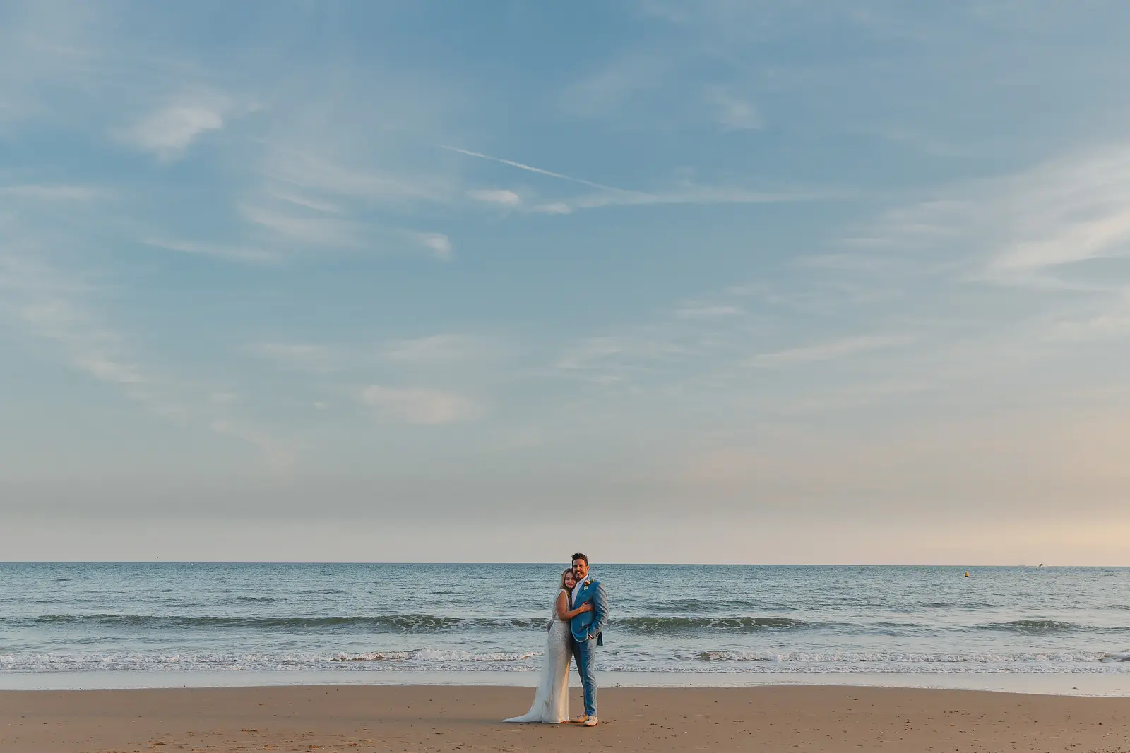 Bride and groom embracing as small figures on a wide sandy Dorset beach at dusk, gentle waves breaking behind them under a pale evening sky