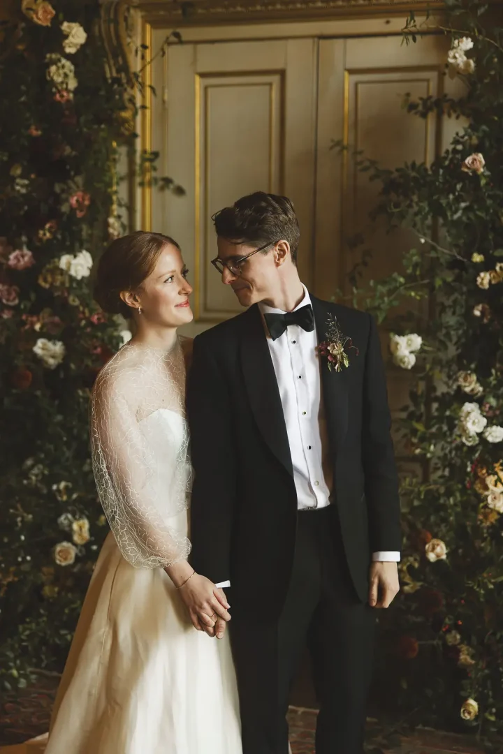 Romantic wedding portrait of bride in net-sleeved gown and groom in black tie looking at each other, framed by a cascading arch of roses in front of a gold-panelled door