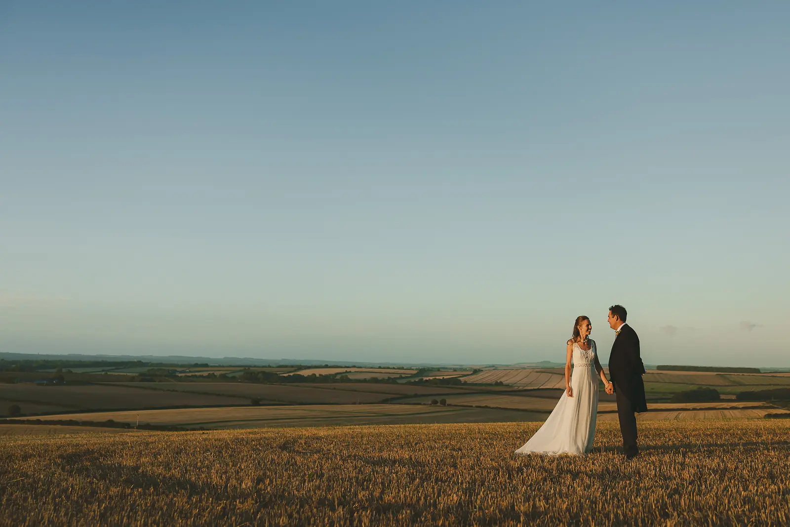 Bride and groom standing hand in hand in a golden wheat field at sunset with a sweeping panorama of the Dorset patchwork countryside, rolling fields and hedgerows stretching to distant hills