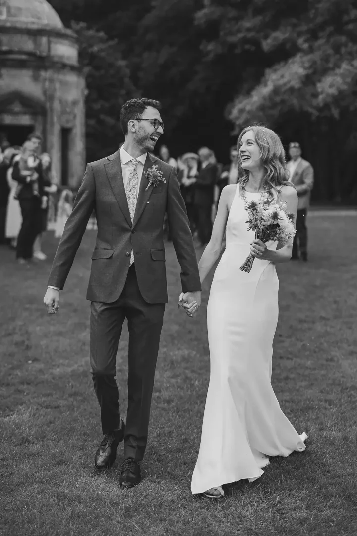 Relaxed black and white wedding portrait of a couple walking hand in hand and laughing across a lawn, a classical stone garden temple visible among mature trees behind them
