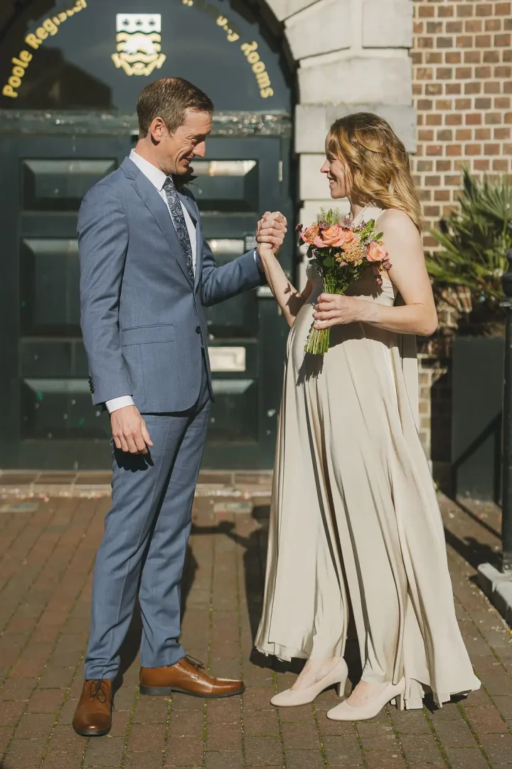 Couple laughing and holding hands outside Poole Registry Rooms entrance, bride in flowing cream dress with coral bouquet