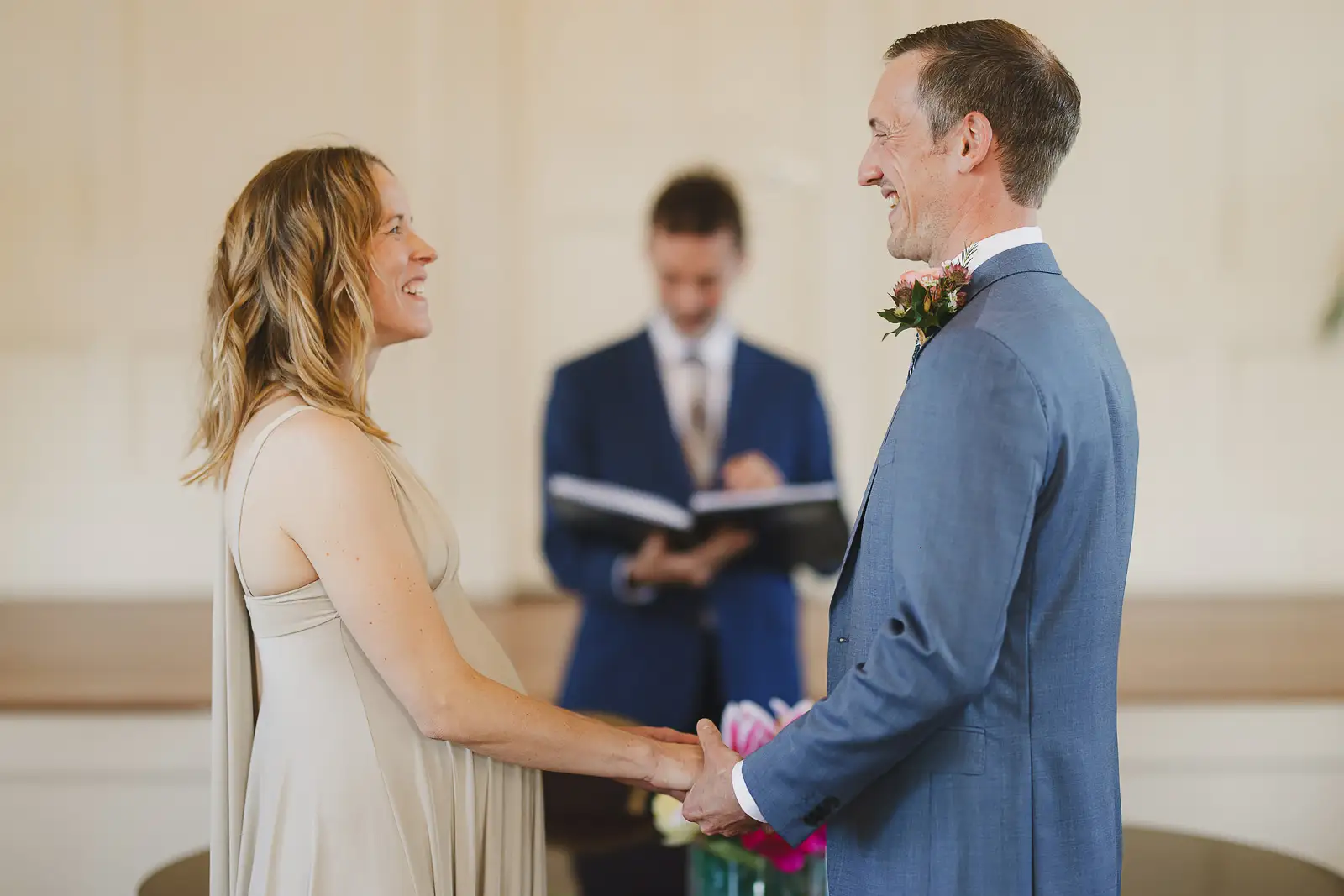 Bride and groom smiling at each other while holding hands during registry office ceremony at Poole Guildhall, registrar behind
