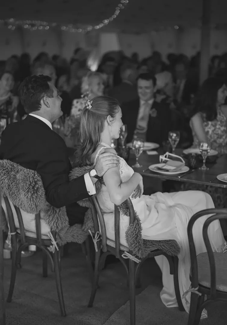 Black and white candid of groom with his arm around the bride's shoulder at the top table during the wedding breakfast in a fairy-lit Dorset marquee, guests seated around them