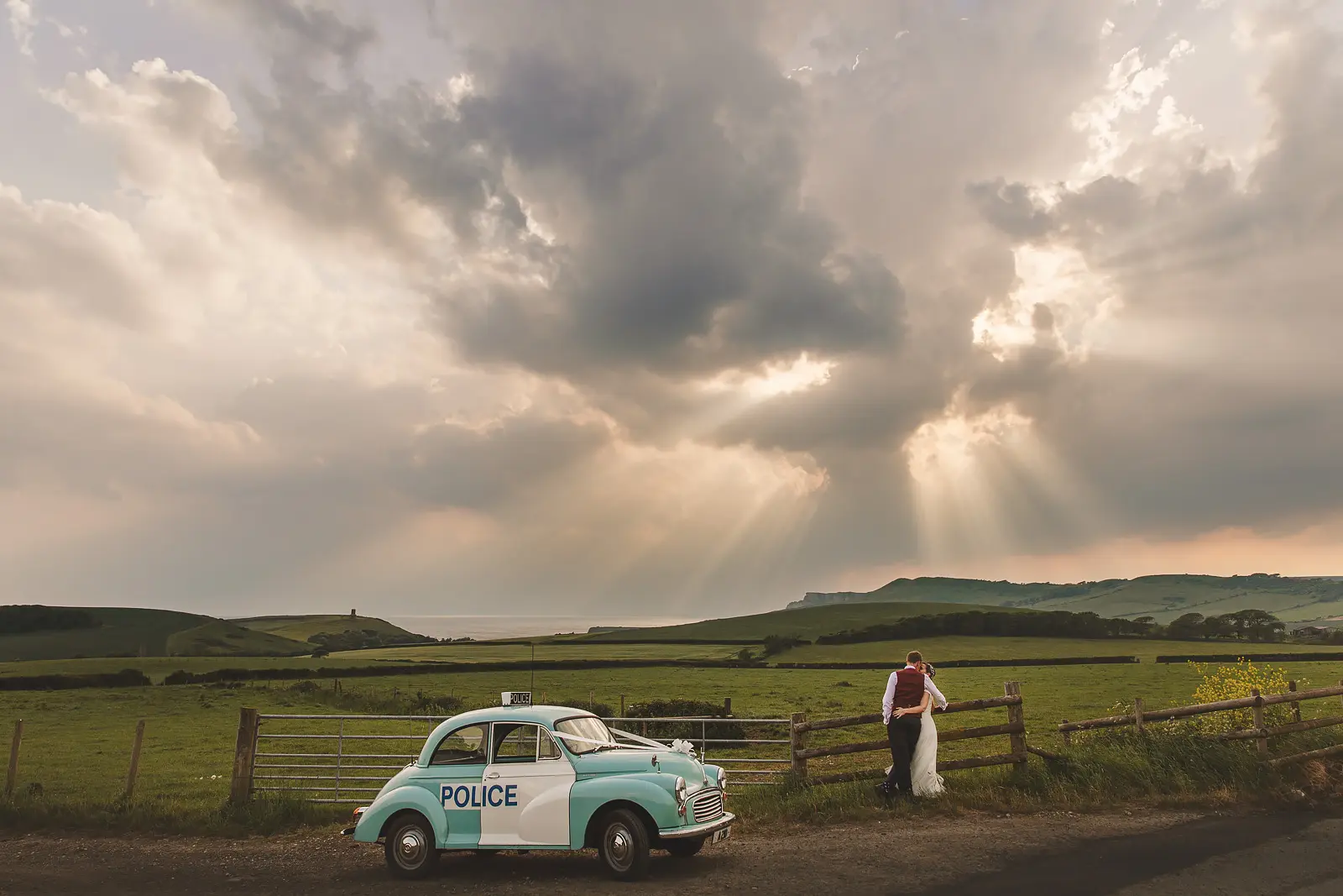 Couple embracing by a fence gate beside a vintage turquoise Morris Minor police car with dramatic sunbeams piercing storm clouds above the rolling Dorset hills and Kimmeridge coastline